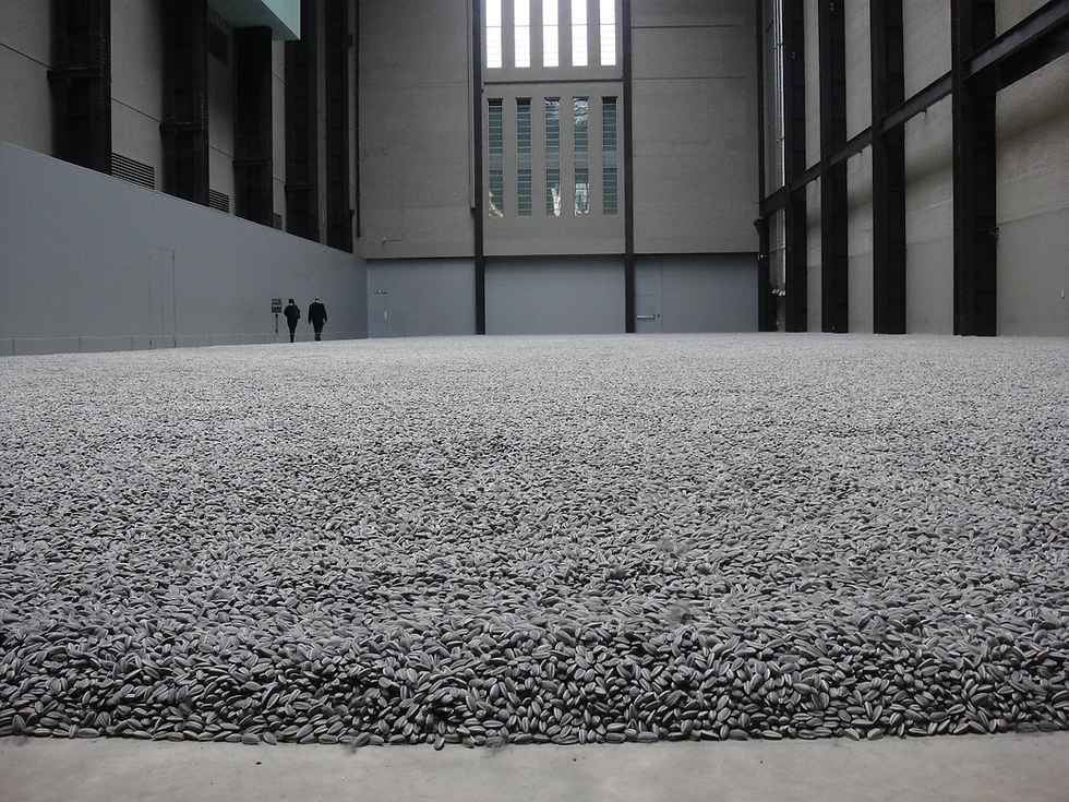 Vast gray hall filled with a carpet of sunflower seeds. Two people walk in the distance, creating a sense of scale and solitude.