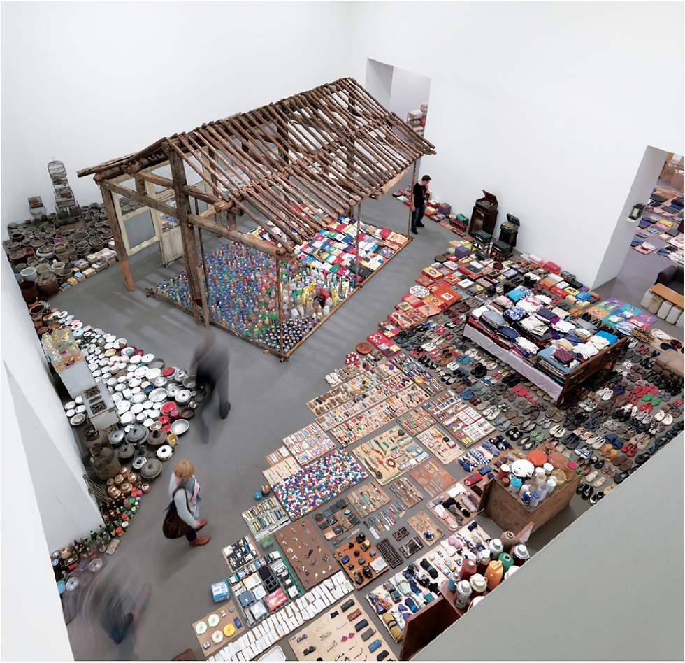 Overhead view of a gallery installation with a rustic wooden hut and neatly arranged household objects, clothes, shoes, and ceramics.