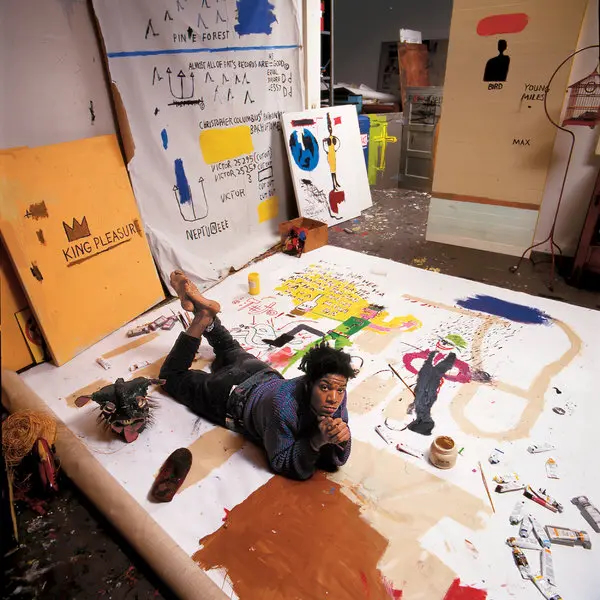 Artist lies on a paint-splattered studio canvas amid colorful works, with KING PLEASURE and PINE FOREST text on panels.