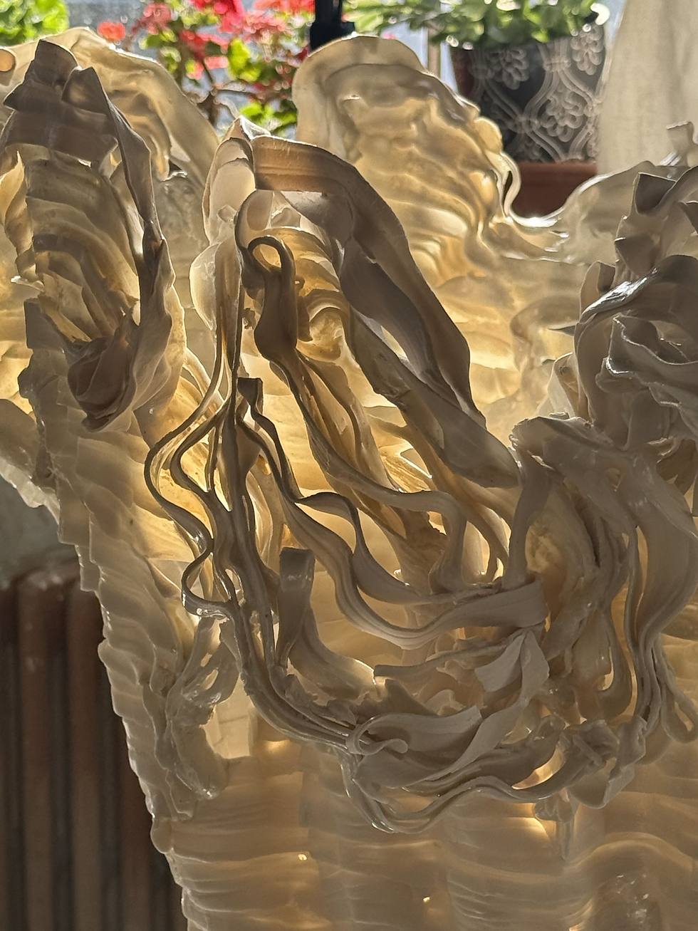 Close-up of a translucent cream glass chandelier with flowing ruffled shapes, backlit by sunlight, with blurred flowers behind.
