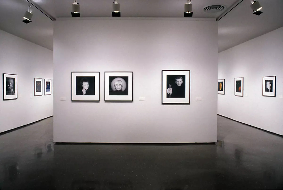 Gallery with black-and-white portraits on white walls, lit by overhead lights. The room is empty, creating a quiet, contemplative mood.