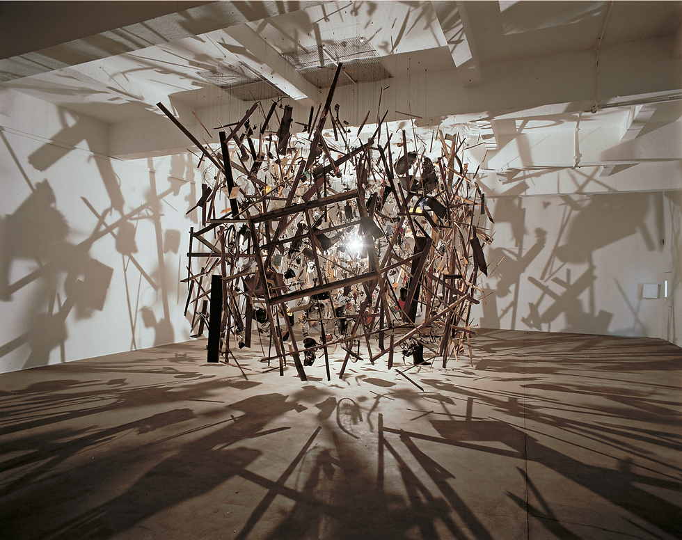 Abstract hanging sculpture of wooden rods in a white gallery, casting dramatic shadows across the floor and walls.