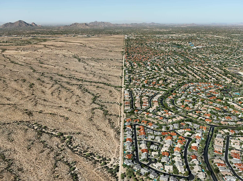 Aerial view of a stark contrast between sprawling desert on the left and dense suburban housing with red roofs on the right. Mountains in the distance.