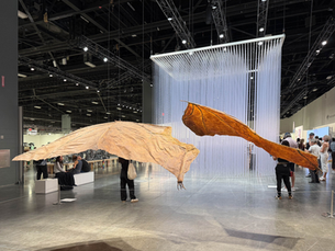Art installation with large, textured wings in a gallery. People observe from a distance. Ceiling beams and hanging lines visible. Industrial setting.