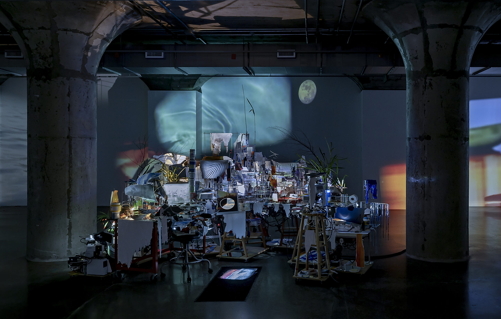 Dim industrial gallery with a cluttered mixed-media installation, projected moonlit imagery, and concrete columns in a moody space