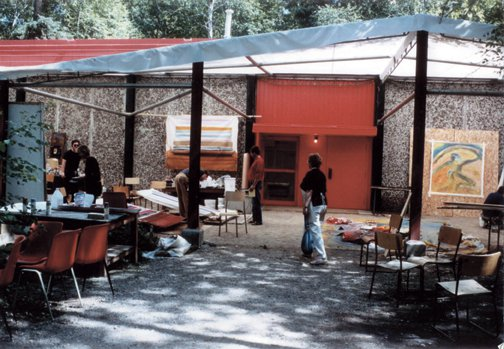 Outdoor art studio with people setting up chairs and art supplies. Two large paintings lean against a red building. Sunny day with trees.