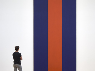 Man in black shirt observes a large abstract painting with vertical blue and orange stripes. Wooden floor reflects the artwork. Museum setting.