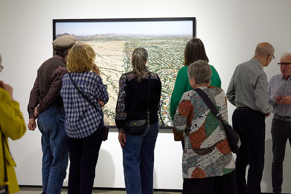 Museum visitors stand before a large aerial landscape photo in a white gallery, quietly studying the framed desert city view.