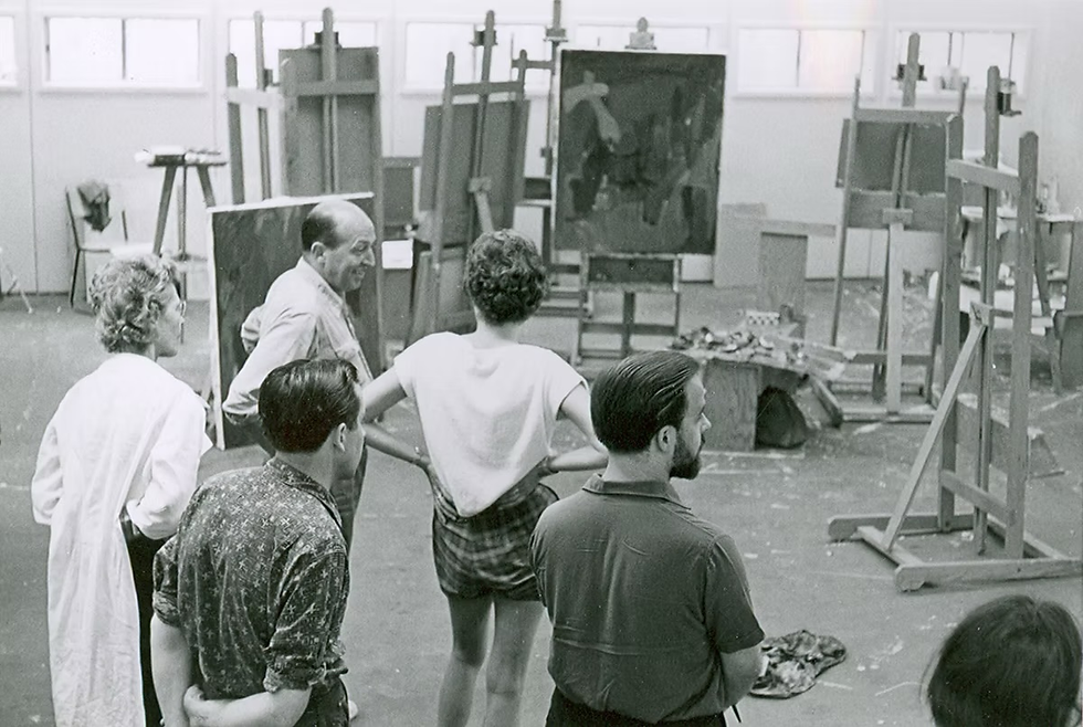 Artists in a studio surrounded by easels and paintings, observing a canvas. The room is filled with natural light, creating a focused mood.