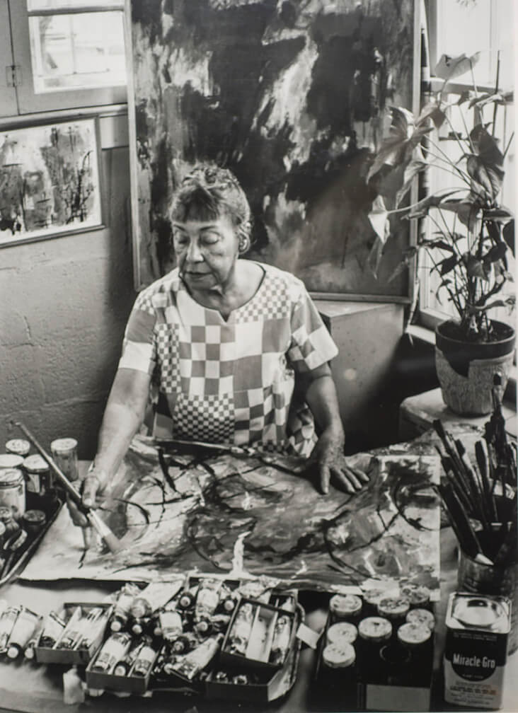 An artist in a checkered shirt paints abstract art in a studio with plants and paint supplies. Black and white photo captures focus and creativity.