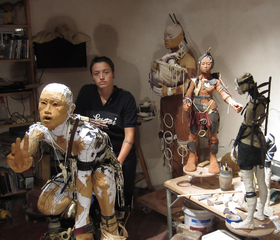 Person in black shirt with diverse sculpture figures in a cluttered studio. The sculptures are detailed with a variety of materials.