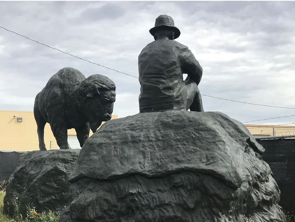 Statues of a person sitting and a bison standing on rocks. Overcast sky with a building and wire fence in the background.