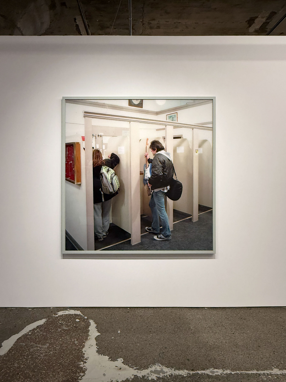 A framed photo on a gallery wall shows two people using phone booths. The setting is indoors with muted colors and a concrete floor.