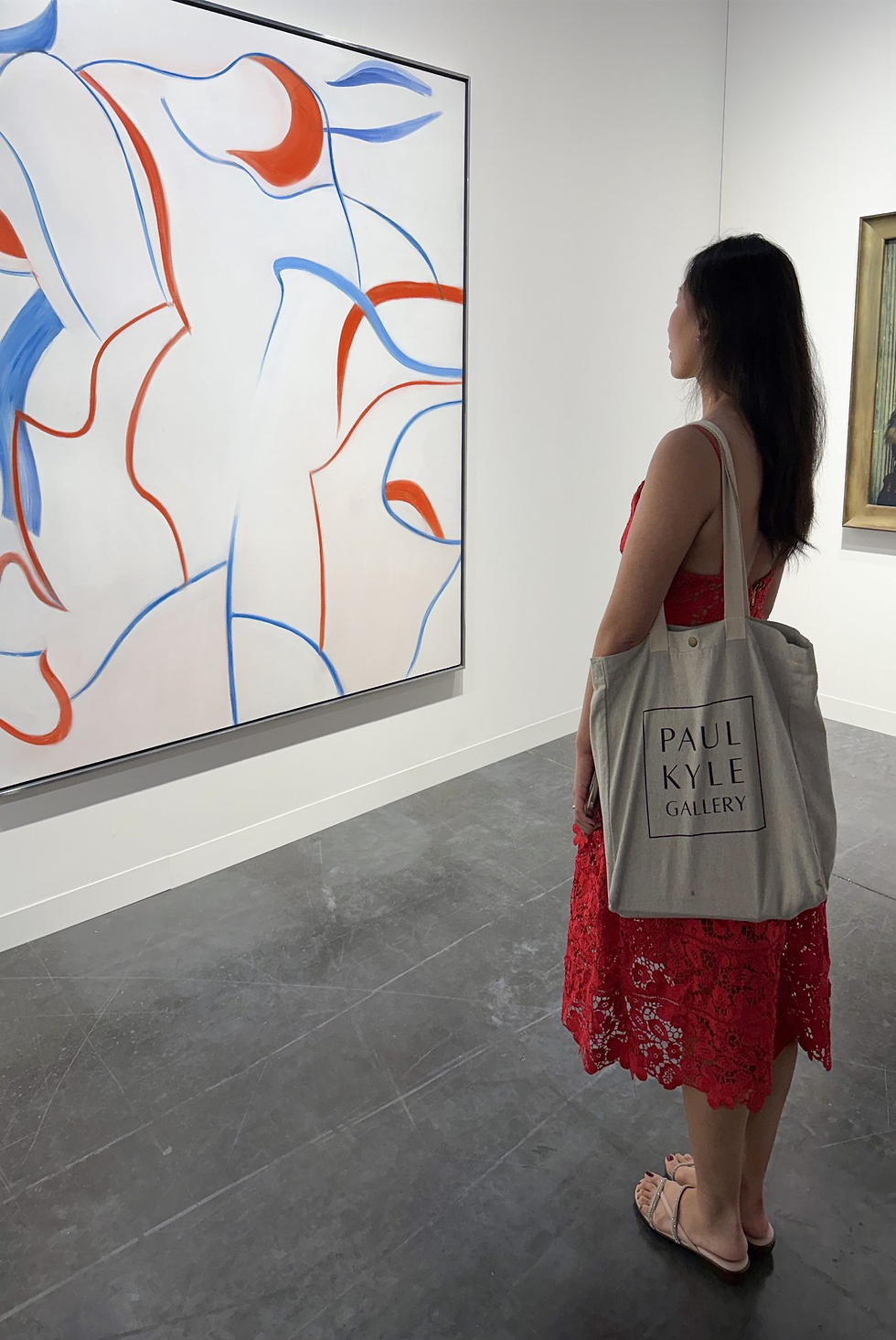 Woman, Diamond Zhou, in red dress admires abstract painting with red and blue lines in gallery. Tote reads "PAUL KYLE GALLERY." Gray floor.