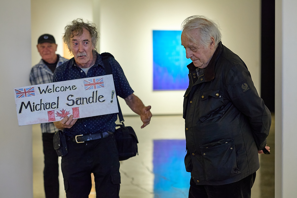 Elderly man holds Welcome Michael Sandle sign with UK and Canada flags as another man looks down in a gallery hallway.