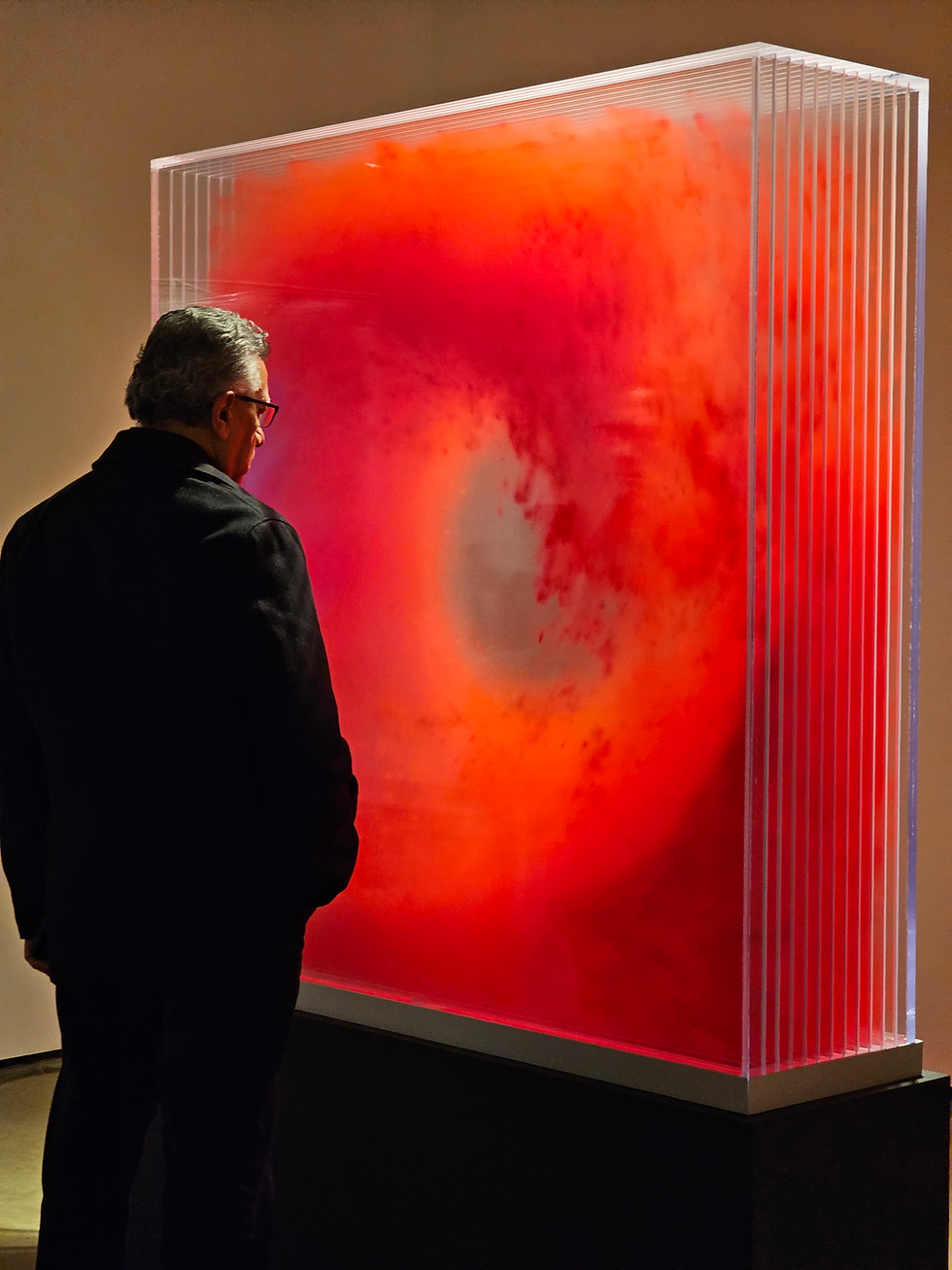 Man in dark coat observes a glowing red and orange abstract art piece in a dimly lit gallery.