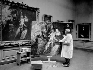 An artist in a white coat paints a religious scene on a large canvas, matching a framed artwork in a gallery. Paints and brushes nearby.