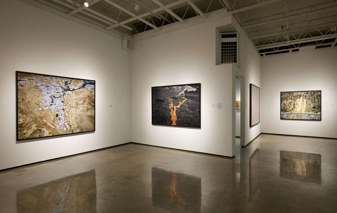 Contemporary art gallery with white walls and polished concrete floors. Large photographs in black frames are installed on the walls in a minimalist aesthetic. Photographs depict an aerial view of slat ponds, the tailings pond of a nickel mine, and a railway cut into a mountainside.