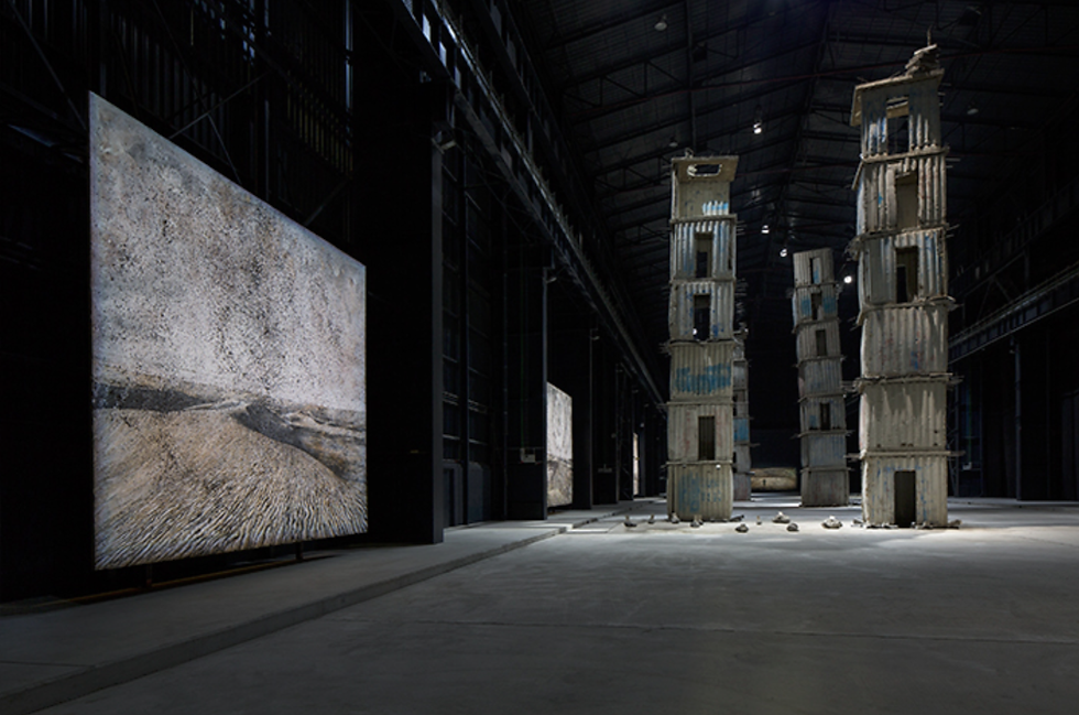 Dim warehouse gallery with a large abstract landscape painting and tall stacked corrugated-metal tower sculptures.