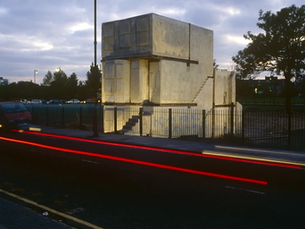 Small weathered two-story house-like building behind a fence at dusk, with red and white car light trails on the road