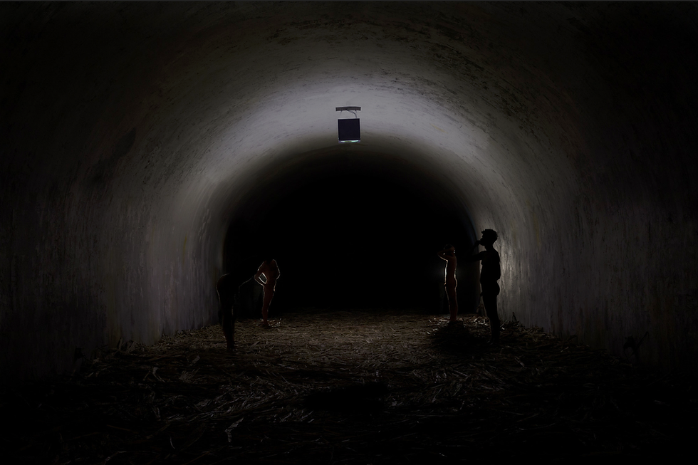 Silhouetted figures stand in a dimly lit tunnel with a light overhead. Moody atmosphere, earthy floor, and shadowy walls.