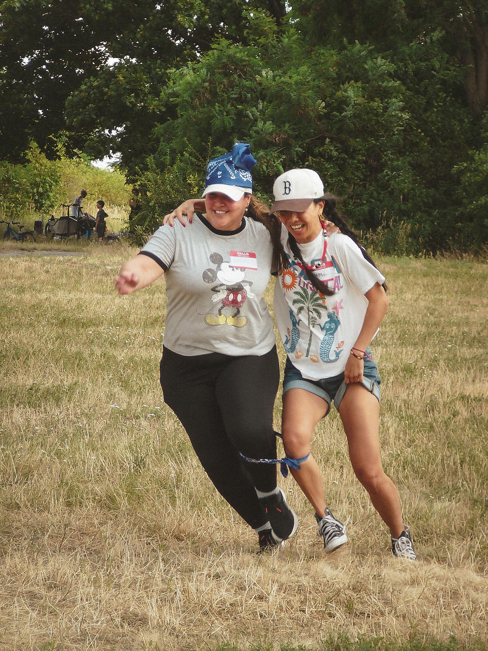 Two smiling women in caps and name tags running a three-legged race at an adult summer camp in Berlin
