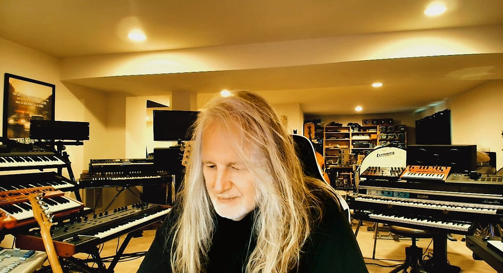 Rick Balentine seated in his home music studio, surrounded by keyboards, synthesizers, and recording equipment.