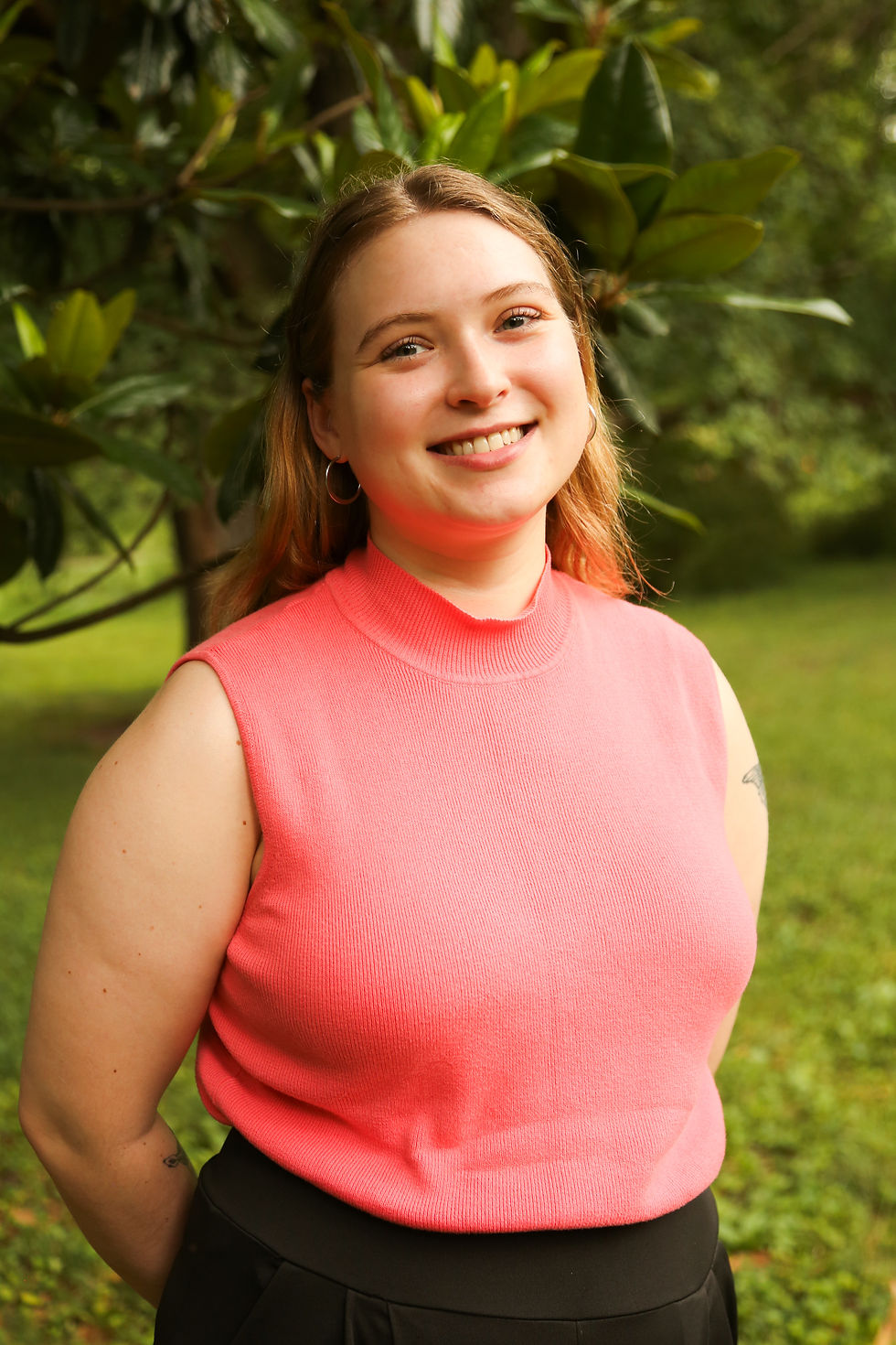 Smiling person in a pink sleeveless top stands before a leafy green background. The mood is cheerful and the scene is outdoors.