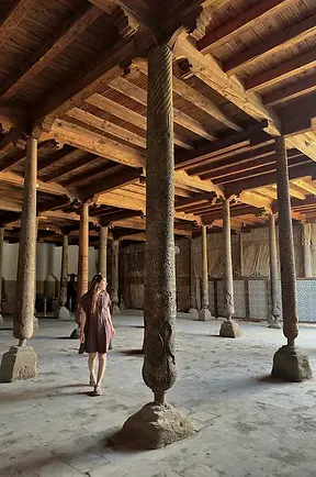 Donna che cammina tra le colonne lignee della moschea Juma di Khiva, spazio religioso storico dell’Asia centrale.