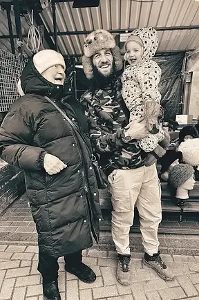 Padre e figlio accanto a un’artigiana russa che realizza colbacchi tradizionali, banco del Mercato Centrale di Riga con tessuti e materiali artigianali.