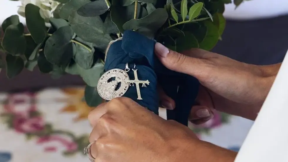 Manos sostienen un ramo de noviacon hojas verdes y blancas, adornado con medallas religiosas sobre un paño azul. Fondo desenfocado colorido.