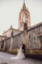 Novios frente a la  catedral  de Oviedo , ella con vestido blanco y ramo, él con traje oscuro. Fondo de piedra, reja y torre alta, ambiente romántico.
