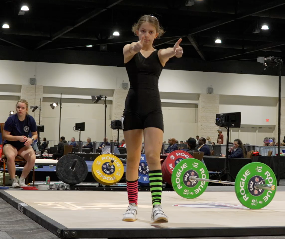 A weightlifter in mixed striped socks gives a thumbs-up on a platform. Barbells and spectators in the background, focused and lively ambiance.