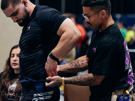 Man in black shirt with number 07 being assisted by another person adjusting his shorts. Background shows a woman sitting and a dark curtain.