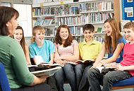 A teacher and a group of six middle-school aged students in a circle with books in a library.