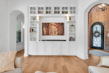 Custom built-in white shelving with glass cabinet doors, integrated lighting, and autumn forest artwork, complemented by warm hardwood flooring and an arched brick entryway in a Lake Austin remodel.