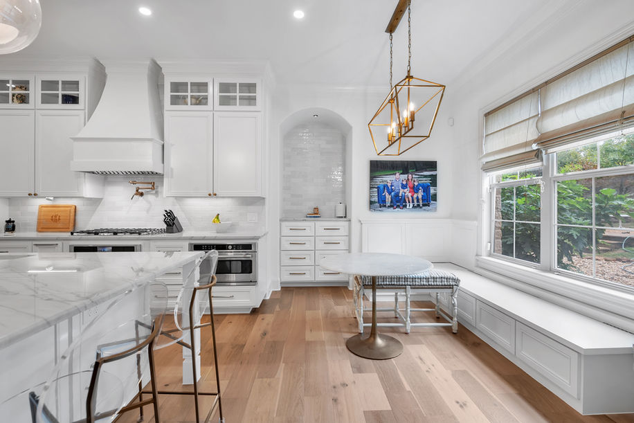 Custom white kitchen with beveled shaker maple cabinets, marble countertops, built-in breakfast nook, gold chandelier, and large windows.