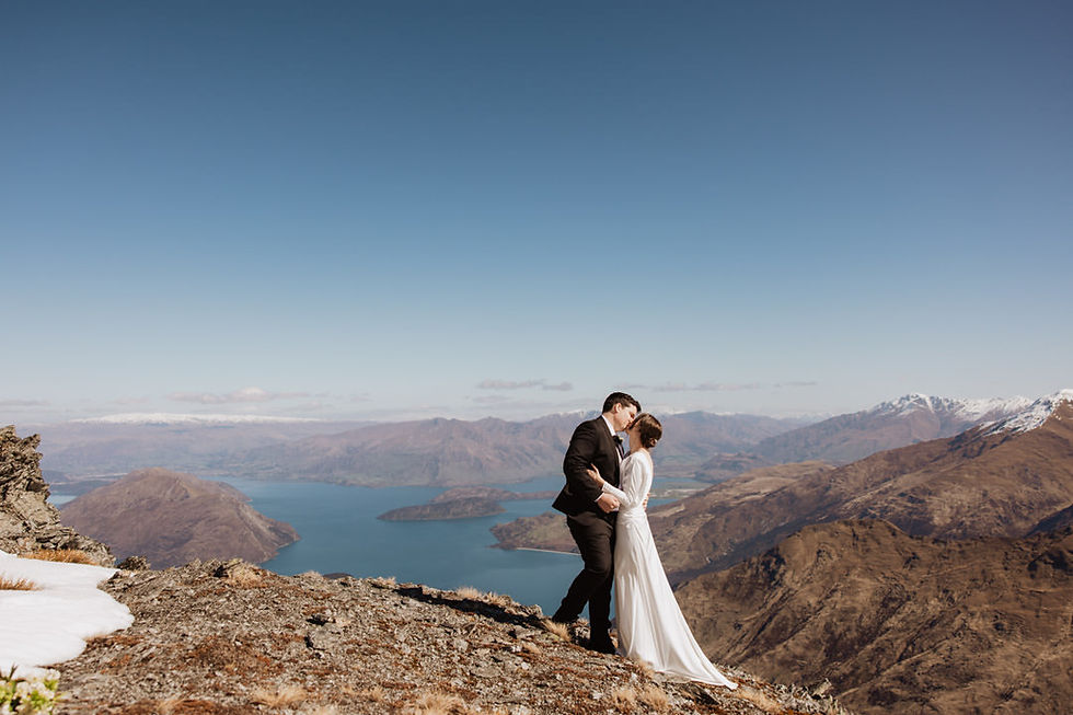 A couple in wedding attire shares a kiss atop a mountain, with a scenic view of a lake and snow-capped peaks under a clear blue sky.