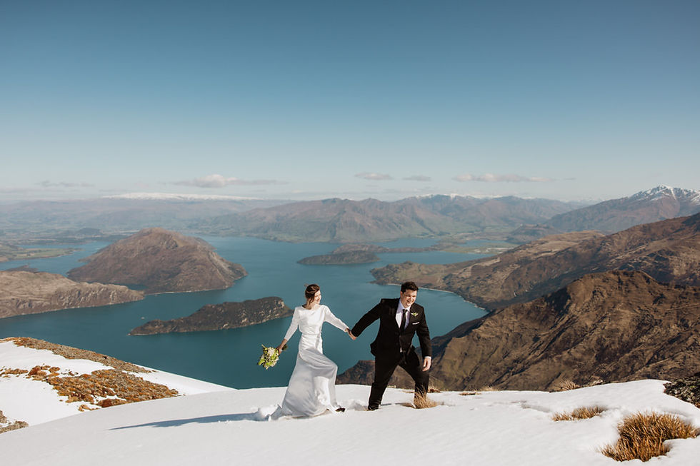 Bride and groom holding hands, walking on snowy mountain with a scenic lake and mountain view in the background under a clear blue sky.