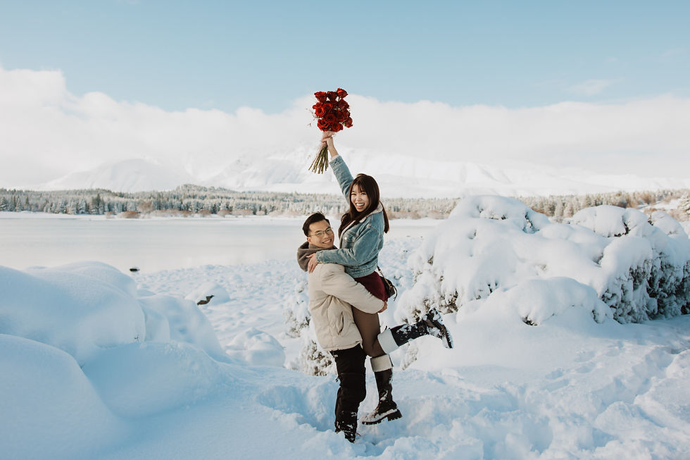 Man lifts a woman holding red roses, smiling in a snowy landscape by a lake. Clear sky and snow-covered bushes create a joyful scene.