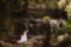 Bride and groom stand smiling by a waterfall in a lush forest, surrounded by greenery and rocks, creating a romantic setting.