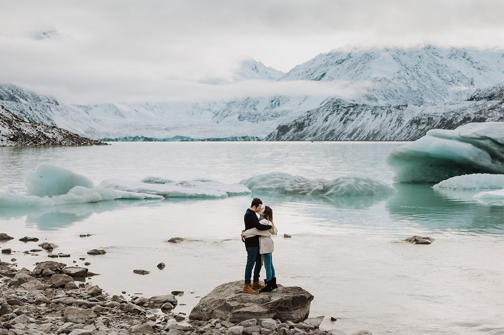 Couple embraces on rocky shore with icy lake and snow-covered mountains in background. Overcast sky, serene mood.