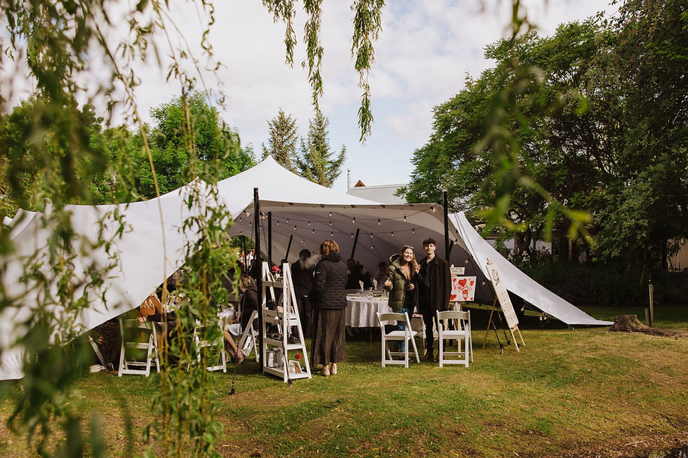 Outdoor event under a white tent with people gathering, tables set, and leafy surroundings. Bright, relaxed atmosphere.