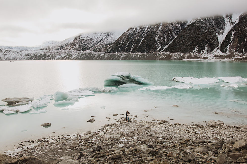 Couple embracing on a rocky shore by a serene, icy lake with floating icebergs. Snow-covered mountains in the background under a cloudy sky.