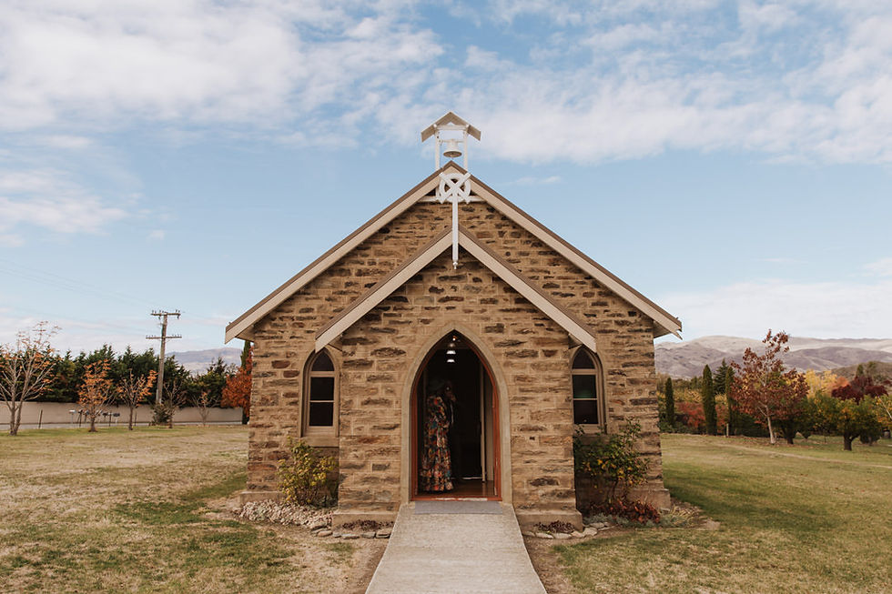 Stone chapel with arched door, surrounded by greenery and autumn trees. Clear sky above; person inside, visible through open door.