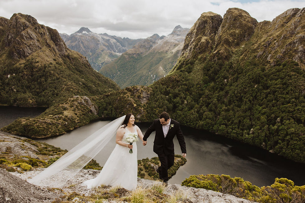 Bride and groom walk hand in hand on a mountain with lake view. The bride wears a white dress, smiling, holding a bouquet. Lush, green scenery.