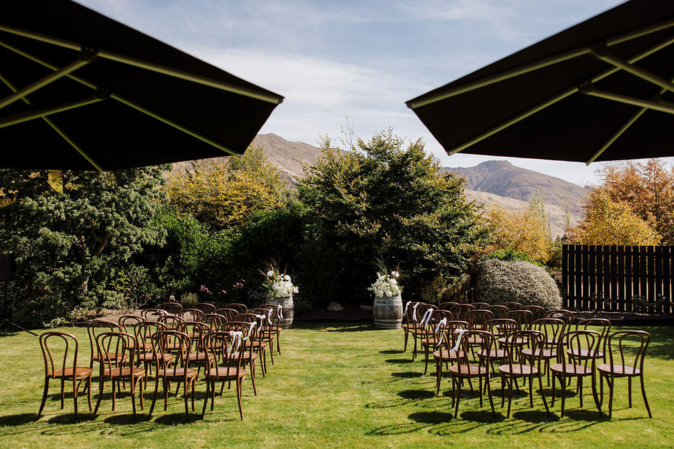 Outdoor wedding setup with wooden chairs in rows, floral arrangements on barrels, under large umbrellas, surrounded by greenery and mountains.