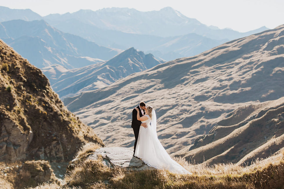 Bride and groom embrace on a mountain peak. Sunlit mountains form the backdrop, creating a serene and romantic atmosphere.