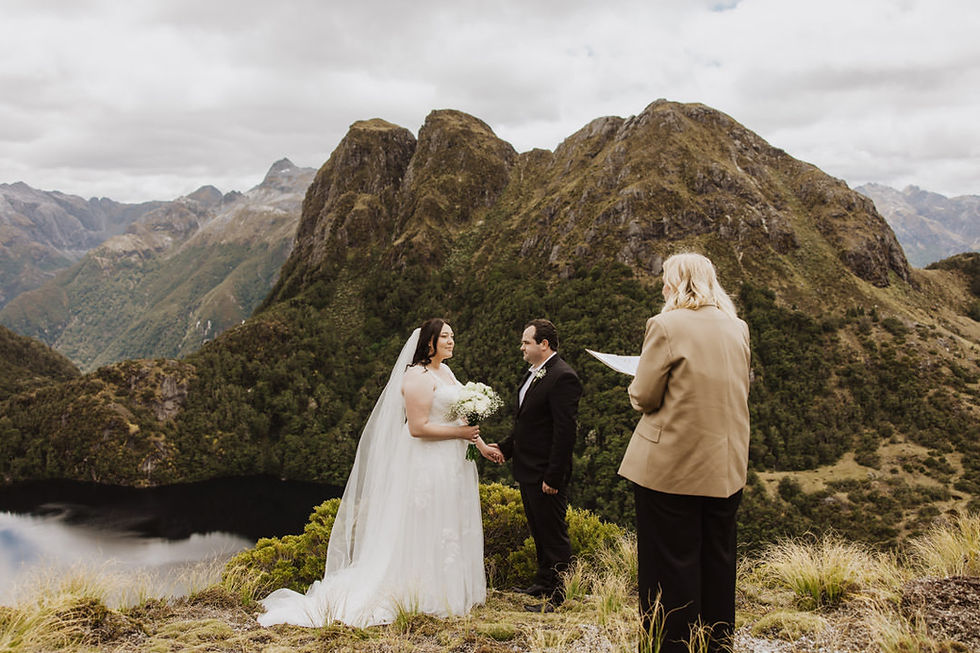 A couple exchanges vows in a mountain setting. Bride in white holds flowers, groom in black suit, officiant reading. Green mountains in background.