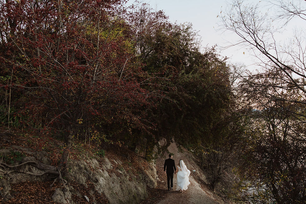 Bride and groom walk down a forest path with lush, overhanging trees. The bride's white dress contrasts with the groom's dark suit. Tranquil mood.